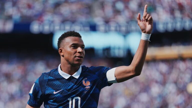 Jeune homme souriant et légèrement perplexe, avec le nom "Kylian Mbappé" en arrière-plan flou, symbolisant l'homonyme face à la célébrité.